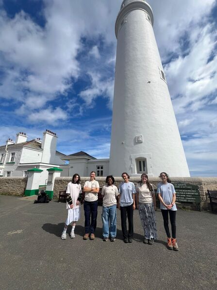 Six young people standing in a line in front of a tall, white lighthouse. Behind the lighthouse is a blue sky with lots of white, fluffy clouds. 
