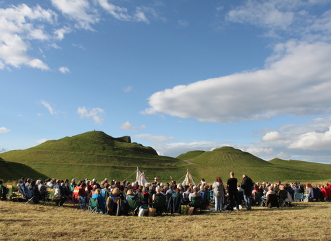 Outdoor theatre event by Handlebards. The audience are sat with their backs to the camera and the actors are in the distance, facing towards the camera. Northumberlandia can be seen in the background.