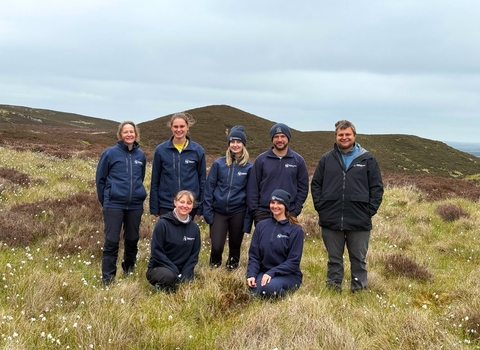 A group of seven individuals in outdoor attire are standing and kneeling in a field of grass and wildflowers against a backdrop of hilly terrain and a cloudy sky.