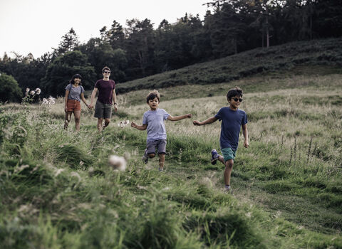 Two children run along a grassy path in open countryside, with two adults walking behind them and a wooded hillside in the background.
