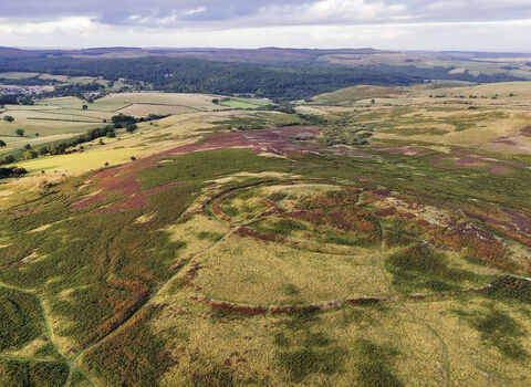 Aerial view of a hill fort with circular earthworks on open moorland, surrounded by rolling fields, woodland, and distant hills beneath a cloudy sky.