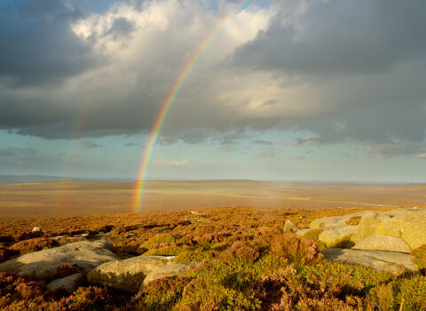 Bright rainbow stretching across a rocky moorland under dramatic dark clouds, with sunlight illuminating the landscape and a faint second rainbow visible in the distance.